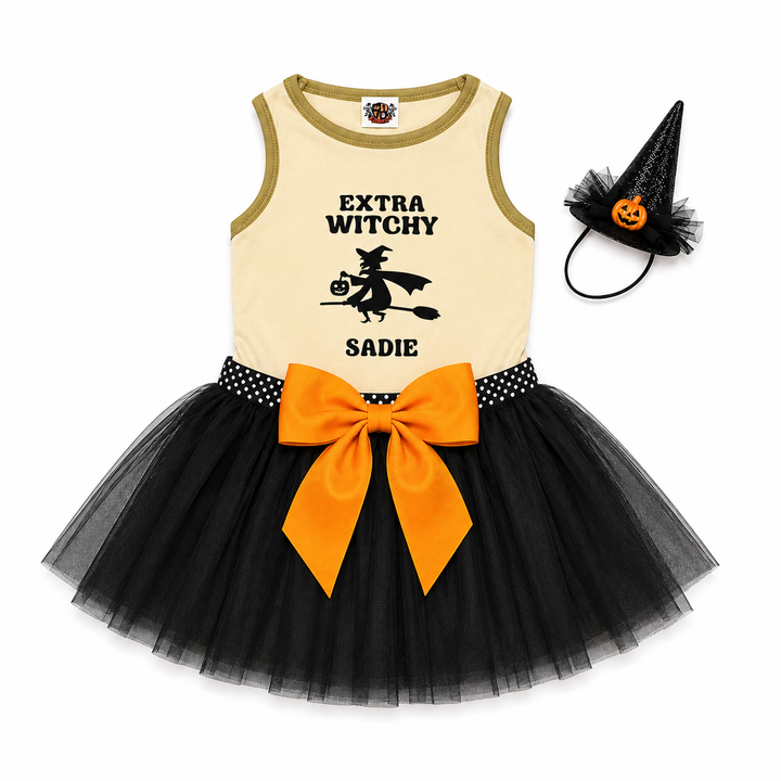 A toddler Halloween costume set featuring a sleeveless cream top with olive trim, a black witch graphic and text, a black polka-dot waistband, a large orange bow, a black tulle tutu skirt, and a matching black pointed witch hat with a jack-o'-lantern face.